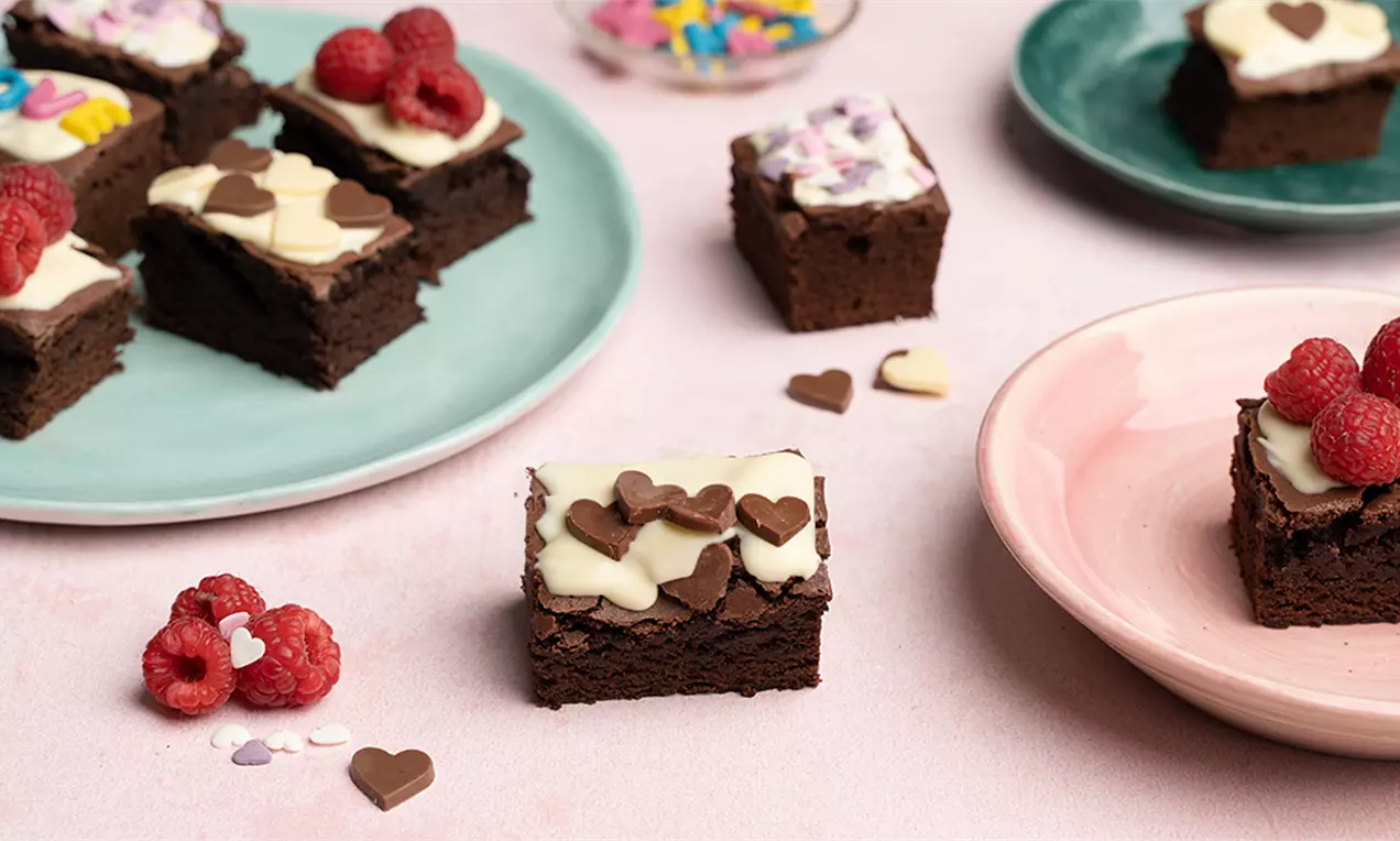 Donkerbruine rechthoekige brownies met witte glazuurlaag, versierd met frambozen, gekleurde letters en hartjes op pastelkleurige borden