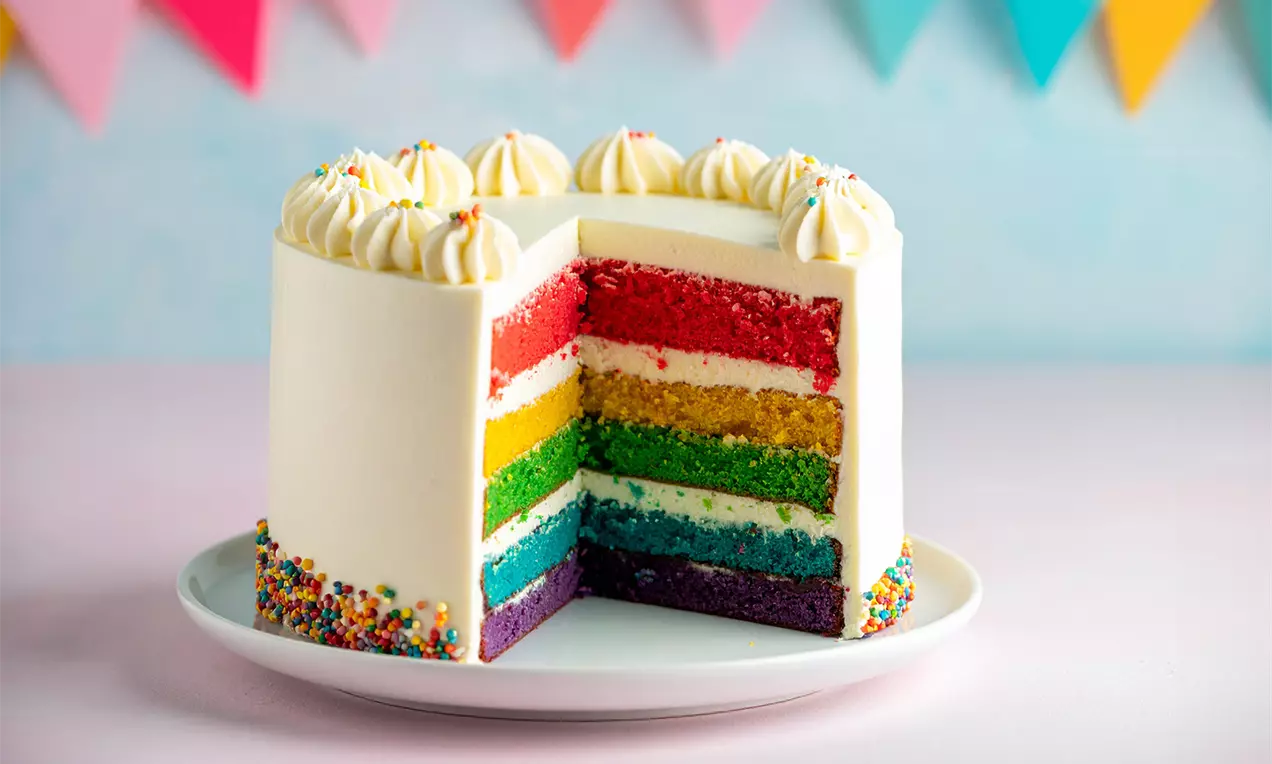 Regenboogtaart met zes gekleurde lagen en witte crème, versierd met toefjes slagroom en regenboogsprinkles op een wit bord