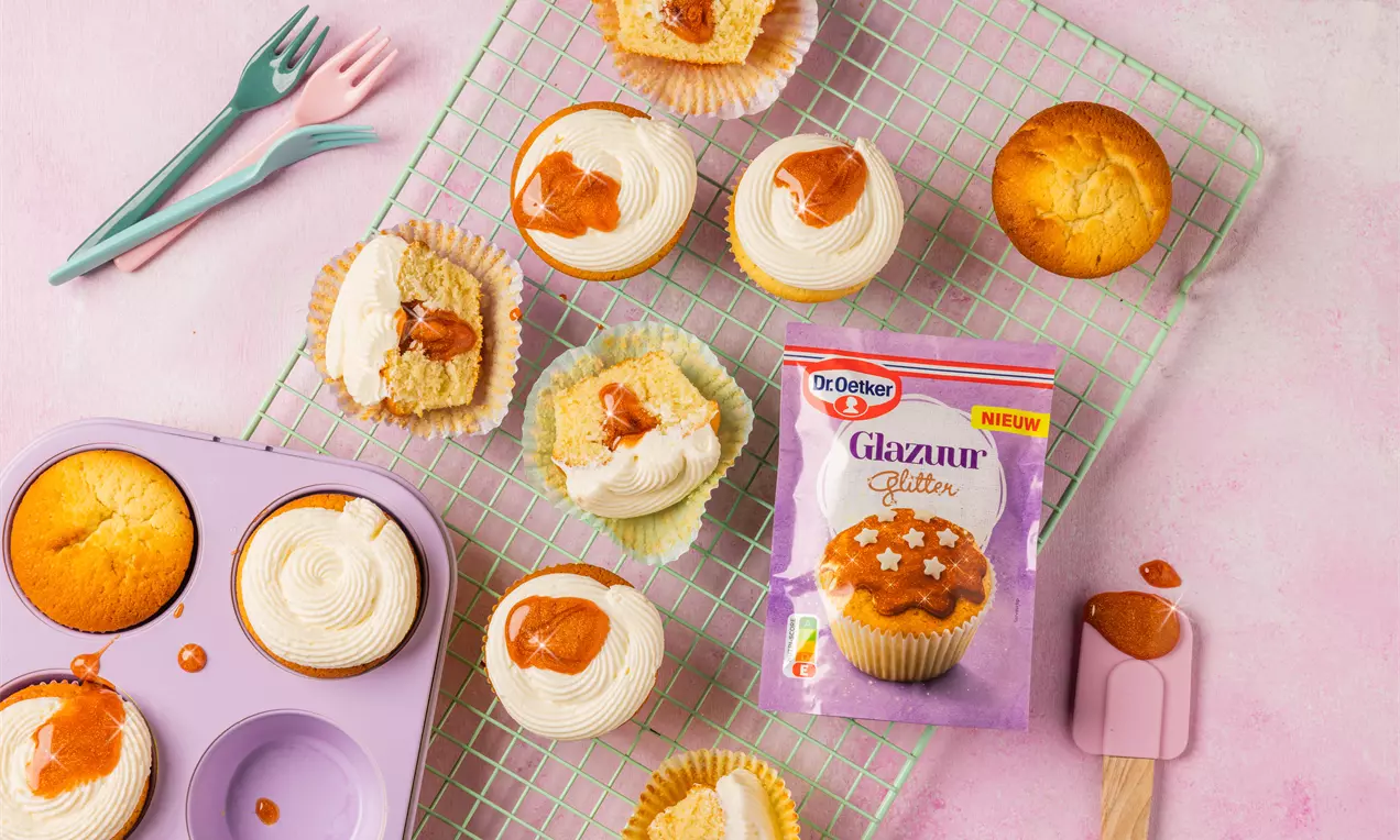 Cupcakes met luchtige cakebodem, opgespoten witte crème en glanzend oranje glazuur, gepresenteerd op een roze ondergrond en een koelrekje