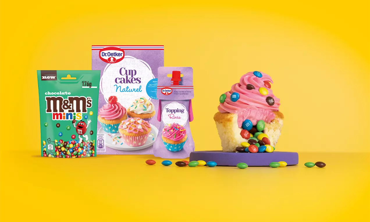 Vanillecupcake met roze glazuur, kleurrijke M&M’s en losse snoepjes op een paarse onderlegger tegen een gele achtergrond