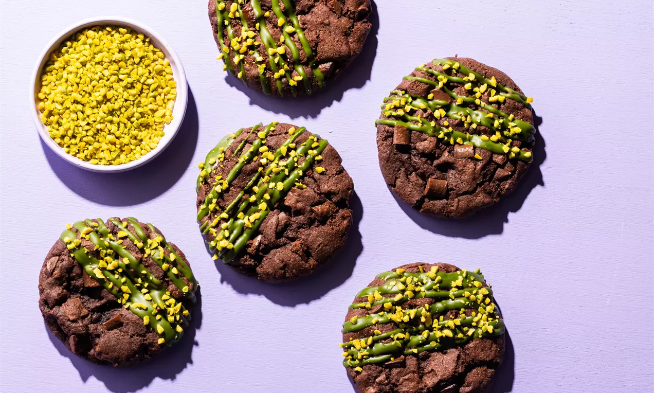 Donkerbruine ronde koekjes met een ruwe textuur, besprenkeld met groene pistachecreme en fijngemalen pistachenootjes op een lila achtergrond